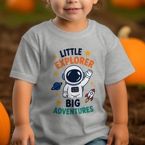 Puede incluir: Una camiseta de manga corta gris jaspeado con un gráfico de un astronauta saludando, un cohete y un planeta. El texto LITTLE EXPLORER BIG ADVENTURES aparece en letras azul oscuro, naranja y verde azulado, con pequeñas estrellas amarillas.