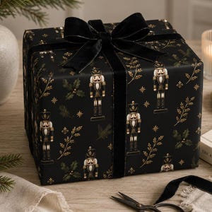 May include: A square gift box wrapped in black paper featuring a repeating pattern of gold nutcrackers, holly leaves, and stars. The box is tied with a wide black velvet ribbon tied in a bow on top, creating a sophisticated and festive presentation.