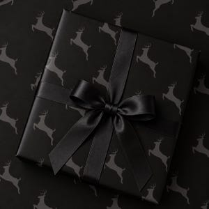May include: A square gift box wrapped in matte black paper featuring a repeating pattern of dark gray leaping deer silhouettes. The box is tied with a wide, smooth black satin ribbon arranged in a centered bow with two long, trailing ends.