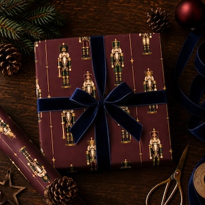 May include: A square gift box wrapped in dark burgundy paper featuring a repeating pattern of nutcracker figures in green and gold uniforms, tied with a wide navy blue velvet ribbon tied in a bow at the center of the package.