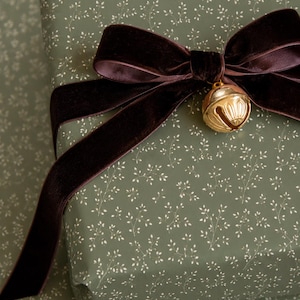 May include: A rectangular gift box wrapped in sage green paper with a delicate white botanical sprig pattern. The package is adorned with a wide, dark brown velvet ribbon tied into a bow, featuring a small, shiny gold-toned metal jingle bell.
