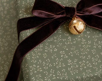 Può includere: Una scatola regalo rettangolare avvolta in carta verde salvia con un delicato motivo a rametti bianchi. Il pacchetto è decorato con un ampio nastro di velluto marrone scuro annodato a fiocco, con un piccolo campanello in metallo dorato.