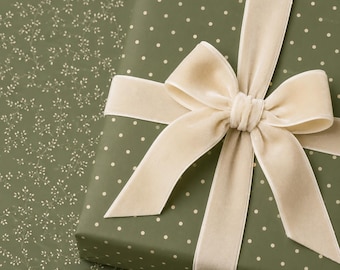 Può includere: Una scatola regalo rettangolare avvolta in carta verde oliva con un motivo a pois bianchi. Il pacchetto è legato con un ampio nastro di velluto color crema, annodato in un grande fiocco a più anelli centrato sulla parte superiore.