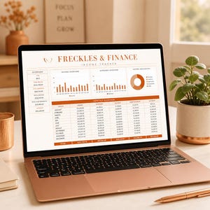 May include: A rose gold laptop displaying a digital financial spreadsheet titled Freckles & Finance Income Tracker. The screen shows an overview, income and expense bar charts, a pie chart, and a monthly table with columns for income, expenses, savings, and debt.