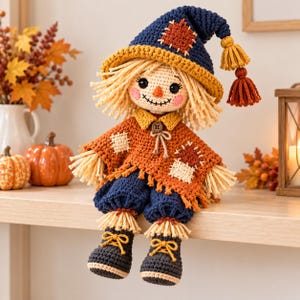 May include: A handcrafted crochet scarecrow doll featuring a navy blue pointed hat with a yellow brim and tassels, a rust-orange poncho with stitched patches, navy blue trousers, and dark boots. The doll has yarn hair and a stitched face.