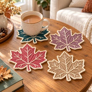 May include: A set of four maple leaf-shaped coasters in teal, purple, red, and tan, each featuring a cream-coloured crocheted border and central vein detailing. One coaster supports a speckled ceramic mug filled with a light brown beverage.