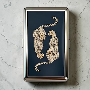 May include: A rectangular metal cigarette case with a polished silver-tone frame and a dark navy blue center panel. The panel features a detailed illustration of two cheetahs in gold and black, positioned facing opposite directions.
