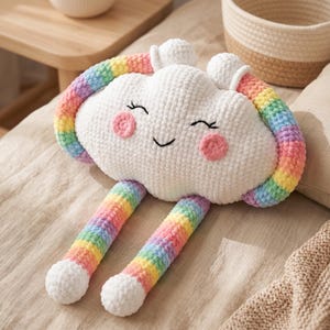 May include: A plush cloud-shaped pillow made of white yarn with a smiling face, pink cheeks, and closed eyes. The pillow features a rainbow-colored yarn border that arches over the top and extends into two long, dangling rainbow-colored legs.