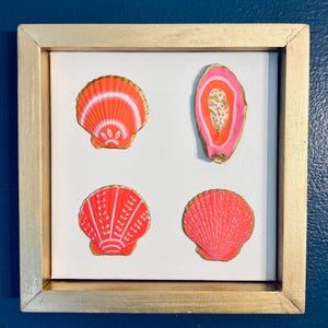 Könnte beinhalten: Ein quadratisches Wandkunstwerk mit vier dekorativen Muscheln, die auf einer weißen Leinwand in einem goldenen Rahmen angeordnet sind. Die Muscheln sind in leuchtenden Neon-Pink- und Orangetönen bemalt, mit goldenen Rändern.