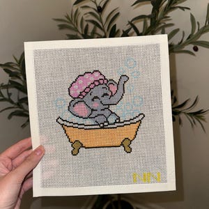 May include: A needlepoint canvas featuring a gray elephant wearing a pink shower cap, sitting in an orange clawfoot bathtub filled with blue bubbles. The design is stitched on a white grid fabric with the yellow letters NN in the bottom corner.