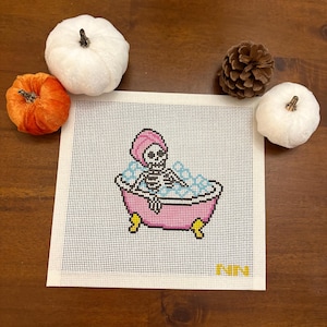 May include: A square needlepoint canvas featuring a skeleton wearing a pink towel on its head, sitting in a pink clawfoot bathtub filled with blue bubbles. The design includes the yellow letters NN in the bottom right corner on a white grid.