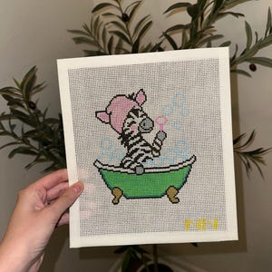 May include: A needlepoint canvas featuring a black and white zebra wearing a pink shower cap, sitting in a green clawfoot bathtub filled with blue bubbles. The zebra is holding a small bubble wand. The letters NN are stitched in yellow at the bottom.