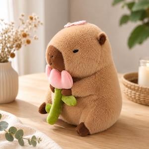 May include: A plush capybara toy with soft light brown fur, a dark brown snout, and closed eyes. The stuffed animal is holding a small, light pink flower with a green stem and leaves, and has a tiny pink flower accessory on its head.