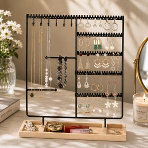 Op de afbeelding: Een zwarte metalen sieradenhouder met een rechthoekige houten voet, voorzien van meerdere haken voor kettingen en armbanden, en geperforeerde stangen voor het ophangen van diverse oorbellen, waaronder ringen, hangers en knopjes.