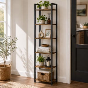 Op de afbeelding: Een hoge, smalle boekenkast met een zwart metalen frame en zes houten planken. De planken zijn gedecoreerd met diverse items, waaronder groene planten in potten, een bruine glazen fles, een beige vaas, een fotolijst, boeken, een kom en een lantaarn.