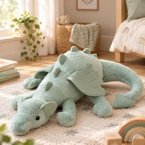 Op de afbeelding: Een zachte, saliegroene pluche draak met een textuurrijke, pluizige buitenkant, kleine vleugels, een lange staart met een puntige uiteinde en een rij kleine stekels op de rug. De draak heeft donkere, ronde ogen en een afgeronde snuit.