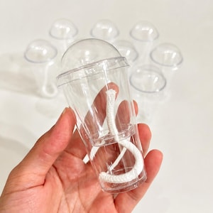 May include: A set of clear plastic containers shaped like miniature cups with dome lids. Each container features a white cotton wick coiled inside the base, designed for candle making. The cups are transparent, lightweight, and stackable.