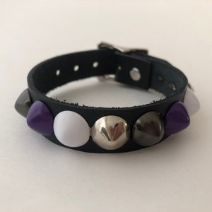Peut inclure: Un bracelet en cuir noir avec des clous argentés, blancs et violets.