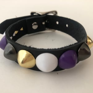 Op de afbeelding: Een zwarte leren armband met zilveren studs en kleurrijke accenten. De armband heeft een zilveren gespsluiting en heeft paarse, witte en gouden studs.