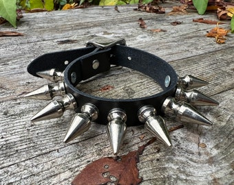 May include: A black leather choker with silver metal spikes. The choker is on a wooden surface.