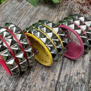 Puede incluir: Tres pulseras de cuero con tachuelas de pir&aacute;mide plateadas. Las pulseras son rojas, amarillas y rosas.