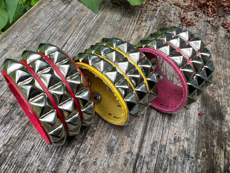 Puede incluir: Tres pulseras de cuero con tachuelas de pir&aacute;mide plateadas. Las pulseras son rojas, amarillas y rosas.