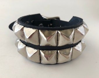 May include: Black leather bracelet with two rows of large, silver pyramid studs. The bracelet has a buckle closure and is shown against a white background. A bold accessory.