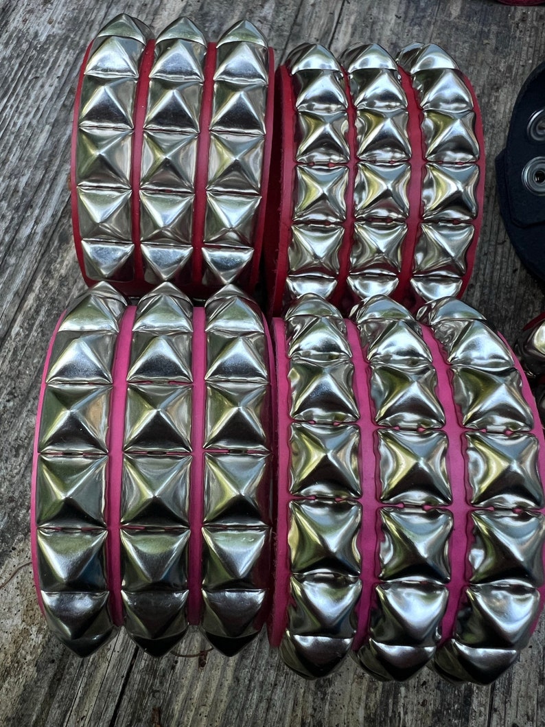 Puede incluir: Cuatro bandas de cuero con tachuelas plateadas y costuras rosas. Las bandas est&aacute;n apiladas una encima de la otra.