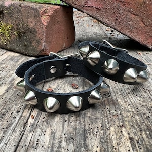 May include: Two black leather bracelets with silver pyramid-shaped studs. The bracelets are on a wooden surface.
