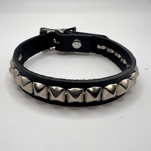 Könnte beinhalten: Schwarzes Lederarmband mit silbernen Pyramiden-Nieten. Das Armband hat einen Schnallenverschluss und einen einzelnen Druckknopf. Die Nieten sind gleichmäßig angeordnet.