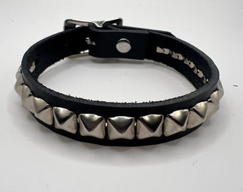 May include: Black leather bracelet with silver pyramid studs. The bracelet has a buckle fastening and a single press stud. The studs are evenly spaced around the bracelet.