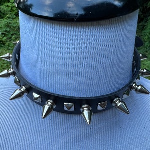 May include: Black leather choker necklace with silver-colored cone and square studs. The choker is displayed on a white mannequin head. The necklace has a buckle closure.