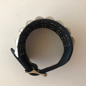 Puede incluir: Un brazalete de cuero negro con tachuelas plateadas y cierre de hebilla.