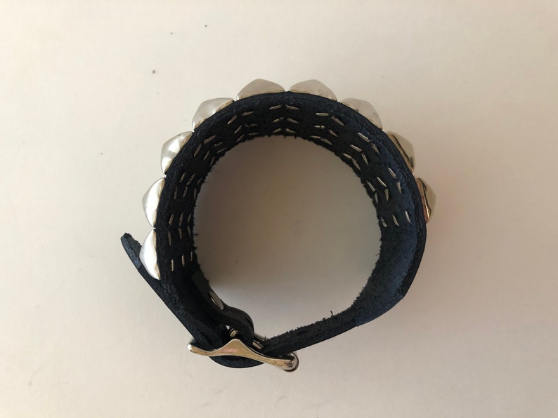 Puede incluir: Un brazalete de cuero negro con tachuelas plateadas y cierre de hebilla.