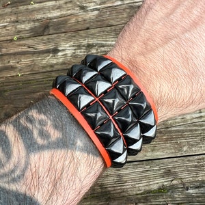 Puede incluir: Un brazalete de cuero negro con una fila de tachuelas en forma de pir&aacute;mide. El brazalete est&aacute; acentuado con una banda de cuero naranja.