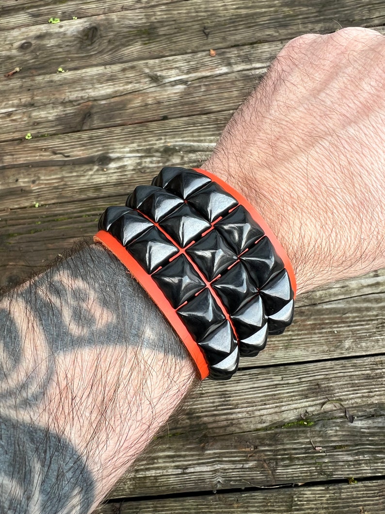 Puede incluir: Un brazalete de cuero negro con una fila de tachuelas en forma de pir&aacute;mide. El brazalete est&aacute; acentuado con una banda de cuero naranja.