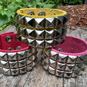 Puede incluir: Tres pulseras de cuero con tachuelas piramidales plateadas. Las pulseras son de color amarillo, rojo y rosa.