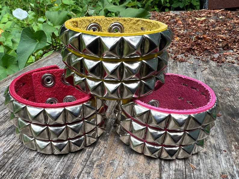 Puede incluir: Tres pulseras de cuero con tachuelas piramidales plateadas. Las pulseras son de color amarillo, rojo y rosa.