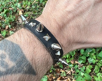 May include: Black leather bracelet with silver-colored cone and star-shaped studs. The bracelet is secured around a wrist. The background is a blurred view of green foliage.