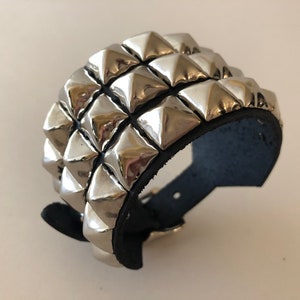 Puede incluir: Un brazalete de cuero negro con tres filas de tachuelas piramidales plateadas.