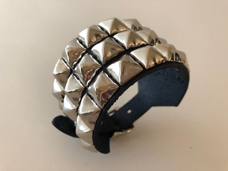 Puede incluir: Un brazalete de cuero negro con tres filas de tachuelas piramidales plateadas.