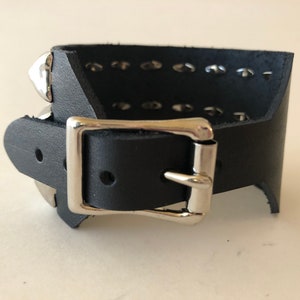 Puede incluir: Pulsera de cuero negro con hebilla plateada y tachuelas piramidales plateadas.