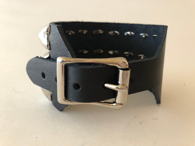 Puede incluir: Pulsera de cuero negro con hebilla plateada y tachuelas piramidales plateadas.