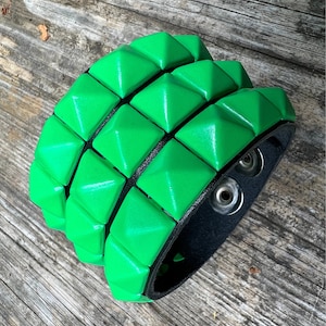 Puede incluir: Un brazalete de cuero verde con una fila de tachuelas en forma de pir&aacute;mide. El brazalete es ajustable con dos broches met&aacute;licos.