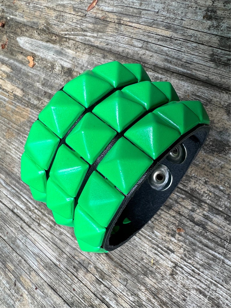 Puede incluir: Un brazalete de cuero verde con una fila de tachuelas en forma de pir&aacute;mide. El brazalete es ajustable con dos broches met&aacute;licos.