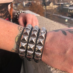 Puede incluir: Un brazalete de cuero negro con tres filas de tachuelas piramidales plateadas.