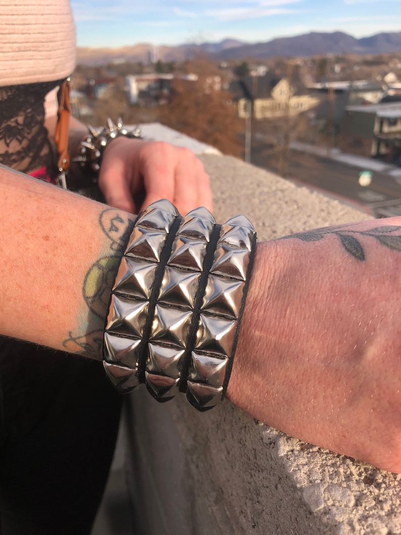 Puede incluir: Un brazalete de cuero negro con tres filas de tachuelas piramidales plateadas.