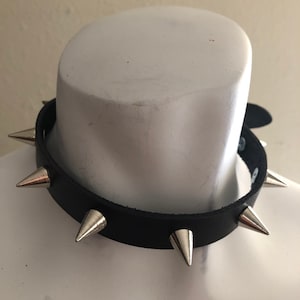 Puede incluir: Un collar de cuero negro con pinchos plateados. El collar está en un maniquí blanco.