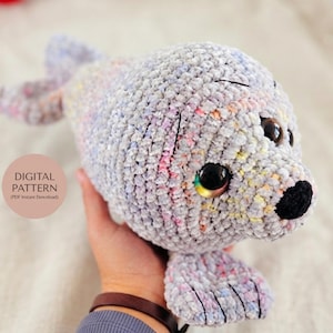 Op de afbeelding: Een gehaakte zeehond knuffel gemaakt van gemêleerd lichtgrijs garen met subtiele pastelkleurige spikkels. Het heeft grote, donkere, glanzende veiligheidsogen, een kleine zwarte geborduurde neus en twee voorvinnen. Een ronde badge toont DIGITAL PATTERN.
