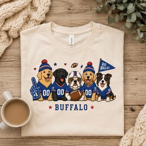 May include: A cream-colored t-shirt featuring a graphic of five dogs wearing blue and red sports jerseys. The dogs are depicted with football accessories including a foam finger, a helmet, a football, and a pennant flag that reads GO BILLS!